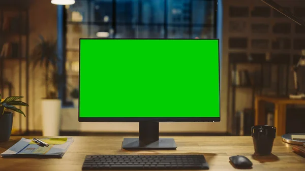 Computador de mesa com tela verde Mock-up em pé na mesa de madeira no escritório criativo moderno. No fundo, iluminação noturna quente e estúdio de espaço aberto com vista para a janela da cidade. — Fotografia de Stock