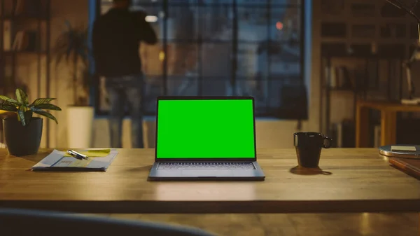 Desktop-Computer mit grünem Bildschirm, der auf dem Holztisch im modernen Kreativbüro steht. Im Hintergrund blickt Designer Drinks aus einer Tasse aus dem Fenster auf die nächtliche Stadt — Stockfoto