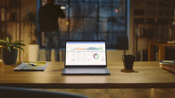 Shot of a Laptop on a Desk with Statistics Graph, Chart and Various Data, Showding Company Growth and Success (англійською). На задньому плані Бізнесмен випиває з чашки і дивиться на Нічне місто з вікна — стокове фото