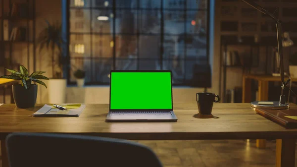 现代创意办公室的办公桌上，立着一个造型精美的绿色屏幕笔记本电脑。城市窗户景观背景下的暖夜照明与开放空间工作室. — 图库照片