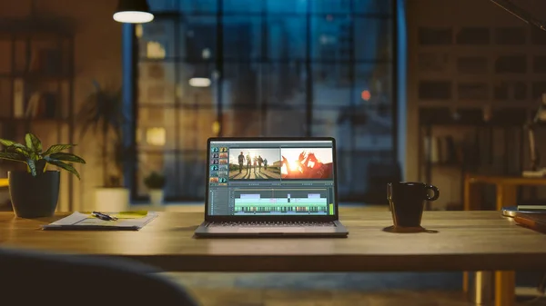 Aufnahme eines Laptops, der auf einem Schreibtisch mit professioneller Videomontage-Bearbeitungssoftware steht. Im Hintergrund warme Abendbeleuchtung und Open Space Studio mit Blick auf die Stadt. — Stockfoto