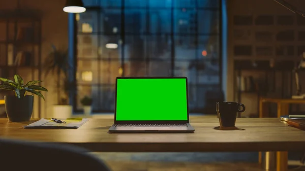 Maquette écran vert ordinateur portable debout sur le bureau dans le bureau de création moderne. En arrière-plan Éclairage du soir chaud et studio à espace ouvert avec vue sur la fenêtre de la ville. — Photo