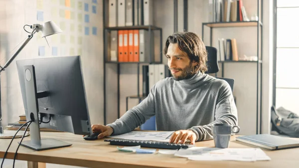 Creative Entrepreneur istuu työpöydällään toimii pöytätietokoneella tyylikkäässä toimistossa. Komea pitkä tukka latino mies taiteellinen näyttää käyttää tietokonetta — kuvapankkivalokuva