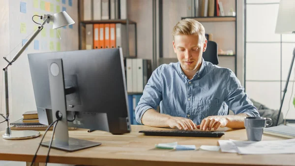 Emprendedor creativo sentado en su escritorio trabaja en la computadora de escritorio en la oficina con estilo. Guapo joven empresario utiliza la computadora, hace outsourcing trabajo, diseña nuevas aplicaciones y desarrolla software —  Fotos de Stock