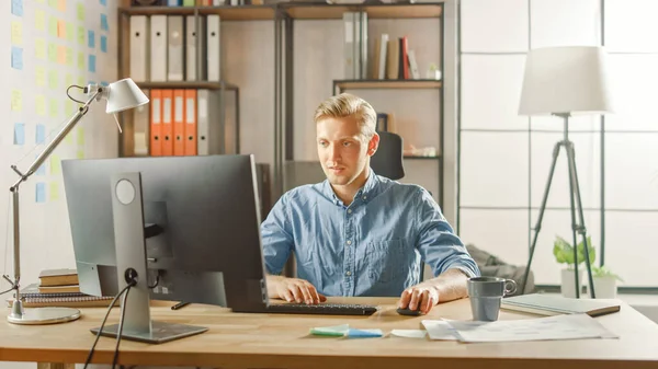 Creative Entrepreneur Sitting at His Desk Works on Desktop Computer in the Stylish Office. Handsome Young Businessman Uses Computer, Does Outsourcing Job, Designs New Apps and Develops Software