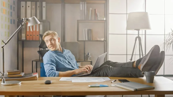 Handsome Young Entrepreneur Works on a Laptop with his legs up on the desk. Designer verwendet Notizbuch, um Software Unicorn Startup zu erstellen. Chill Student Schreibpapier für die Universität — Stockfoto