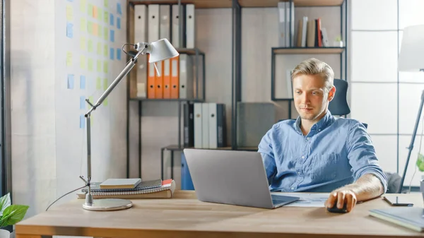 Creative Entrepreneur Sitting at His Desk Works on a Laptop in the Stylish Office, Uses Notebook for Social Media Apps, Emailing Business Associates, Reading News, Browsing through Internet