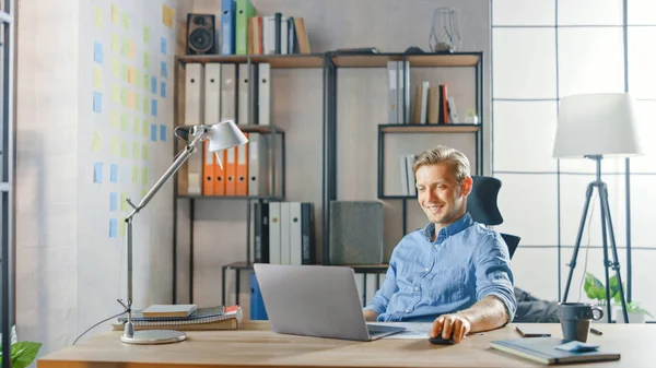 Creative Entrepreneur Sitting at His Desk Works on a Laptop in the Stylish Office, Uses Notebook for Social Media Apps, Emailing Business Associates, Reading News, Browsing through Internet