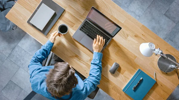 Dans le bureau Un homme d'affaires assis au bureau en bois travaille sur un ordinateur portable dans son bureau à domicile. Il boit du café, des types, écrit des courriels, surfe sur Internet, conçoit des logiciels. Vue du dessus — Photo