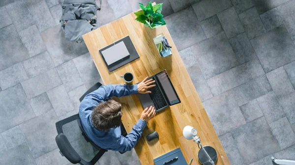 Aufnahme von oben: Geschäftsmann sitzt am Holztisch und arbeitet an einem Laptop in seinem Home Office. Er tippt, schreibt, surft im Internet, erstellt und entwirft Software, Online-Shopping — Stockfoto