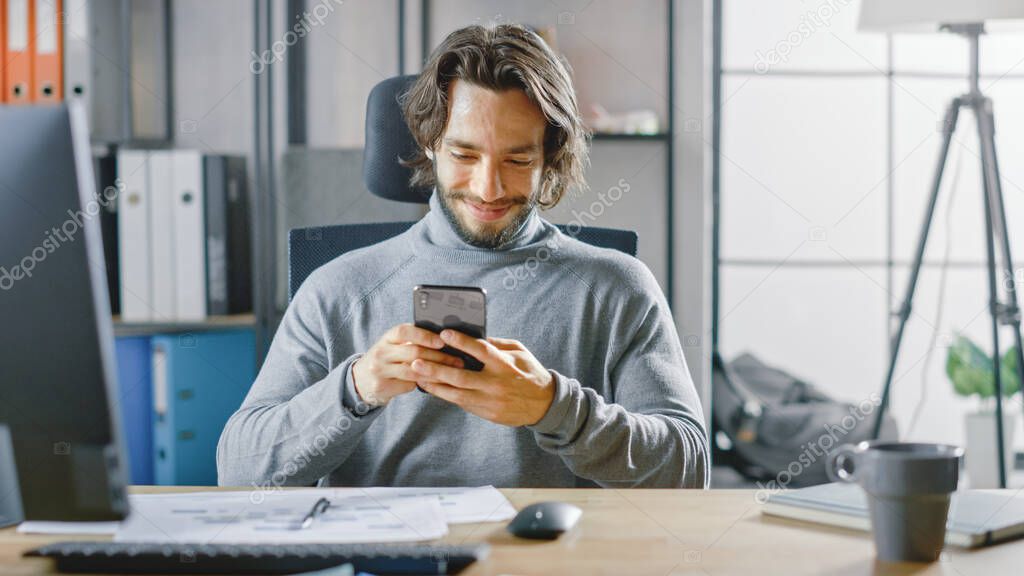 Handsome Long Haired Entrepreneur Sitting at His Desk in the Office Works on Desktop Computer, Working with Documents, Graphs. Uses Smartphone, Social Media App, Writing Emails, Messaging