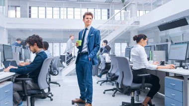 Genç Mutlu Yakışıklı Menajer Takım Elbise Giyiyor ve Uzun Tahtaya Kravat Sürüyor. Elinde bir fincan kahve tutuyor. Modern Açık Ofiste Çeşitli ve Motivasyonlu İş Adamları Bilgisayarlar Üzerinde Çalışıyor.