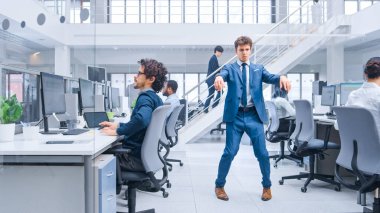 Genç Neşeli Yakışıklı İş Müdürü Ofiste Takım Elbise Giyip Kravat Dansı Yapıyor. Robot gibi dans ediyor. Modern Açık Ofiste Çeşitli ve Motivasyonlu İş Adamları Bilgisayarlar Üzerinde Çalışıyor.