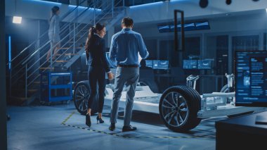 Otomobil Endüstrisi Tasarım Tesisi: Erkek Baş Mühendis Araba Prototipini Kadın Araba Tasarımcısına Gösteriyor. Elektrikli Araç Platformu Chassis Kavramının Tekerlekleri, Motoru ve Bataryası Var. Ekranlarda 3B Cad Yazılımı Göster