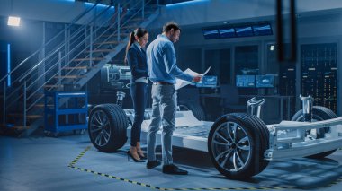 Otomotiv Endüstrisi Tasarım Tesisi: Erkek Mühendis Otomobil Planlarını Gösteriyor Kadın Yazılım Tasarım ve Bütünleşme Mühendisi. Elektrikli Araç Platformu Chassis Konsepti Tekerlekleri, Motoru ve Bataryası Var 