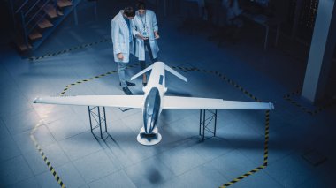 İki Uzay Mühendisi İnsansız Hava Aracı Drone Prototipi üzerinde çalışıyor. Beyaz Önlüklü Havacılık Bilimadamları konuşuyor, Tablet Bilgisayar kullanıyor. Gözetleme ya da Askeri Uçak Endüstriyel Laboratuvarı