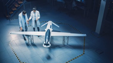 İki Uzay Mühendisi İnsansız Hava Aracı Drone Prototipi üzerinde çalışıyor. Beyaz Önlüklü Havacılık Bilimadamları konuşuyor, Tablet Bilgisayar kullanıyor. Gözetleme ya da Askeri Uçak Endüstriyel Laboratuvarı