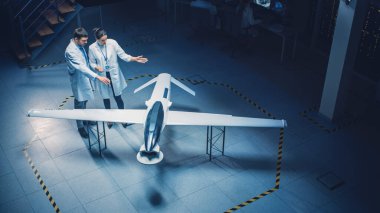 İki Uzay Mühendisi İnsansız Hava Aracı Drone Prototipi üzerinde çalışıyor. Beyaz Önlüklü Havacılık Bilimadamları konuşuyor, Tablet Bilgisayar kullanıyor. Gözetleme ya da Askeri Uçak Endüstriyel Laboratuvarı