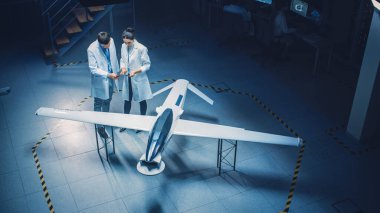 İki Uzay Mühendisi İnsansız Hava Aracı Drone Prototipi üzerinde çalışıyor. Beyaz Önlüklü Havacılık Bilimadamları konuşuyor, Tablet Bilgisayar kullanıyor. Gözetleme ya da Askeri Uçak Endüstriyel Laboratuvarı