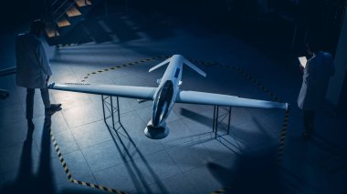 İki Uzay Mühendisi İnsansız Hava Aracı Drone Prototipi üzerinde çalışıyor. Havacılık bilimcileri Tablet Bilgisayar kullanarak konuşuyor. Gözetleme Askeri Uçağı Endüstriyel Laboratuvarı. Yükseltilmiş Vuruş