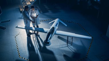İki Uzay Mühendisi İnsansız Hava Aracı Drone Prototipi üzerinde çalışıyor. Beyaz Önlüklü Havacılık Bilimadamları konuşuyor, Tablet Bilgisayar kullanıyor. Gözetleme Askeri Uçaklı Endüstriyel Laboratuvar