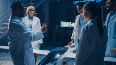 İnsansız Hava Aracı Drone Prototipi üzerinde çalışan Uzay Mühendisleri Toplantısı. Beyaz önlüklü havacılık bilimcileri konuşuyor. Endüstriyel Laboratuvarda Ticari Hava Gözetleme Uçağı