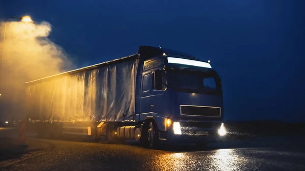 蓝色长沙漏半车，载满货物拖架，夜间旅行，在高速公路上转弯，在雨中驾车穿越大陆，雾雪。工业仓库区. — 图库照片