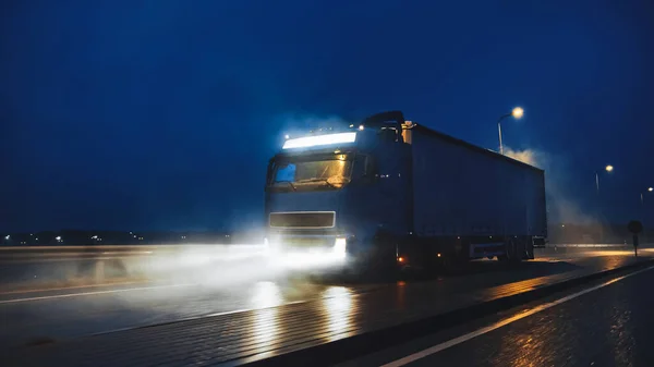 Semi-camion long-courrier bleu avec remorque pleine de marchandises voyage la nuit sur la route autoroutière, traversant le continent à travers la pluie, le brouillard, la neige. Zone des entrepôts industriels. Tir frontal — Photo