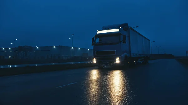 Semi-camion long-courrier bleu avec remorque pleine de marchandises voyage la nuit sur la route autoroutière, traversant le continent à travers la pluie, le brouillard, la neige. Zone des entrepôts industriels. Tir frontal — Photo
