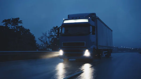 Semi-camion long-courrier avec remorque pleine de marchandises voyage la nuit sur la route autoroutière, traversant le continent à travers la pluie, le brouillard, la neige. Zone des entrepôts industriels. Tir frontal — Photo
