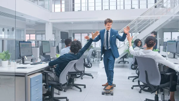 Ung glad stilig chef bär kostym och slips rider en Longboard. Ge high five till kollegor. Mångfaldiga och motiverade affärsmän arbetar med datorer i moderna öppna kontor. — Stockfoto