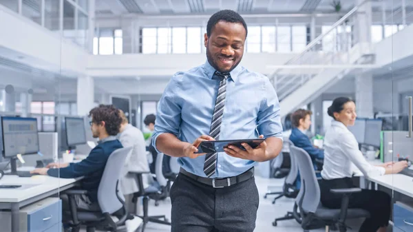 Handsome Young Black Manager in a Shirt Walking Pass His Business Colleagues met een tablet en superviseren hun werk. Diverse en gemotiveerde zakenmensen werken aan computers in de moderne open kantooromgeving. — Stockfoto