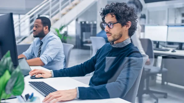 Young Handsome Manager com cabelo encaracolado funciona em um computador desktop e compara dados com um software. Pessoas de Negócios Diversos e Motivados Trabalham em Computadores no Open Office Moderno. — Fotografia de Stock