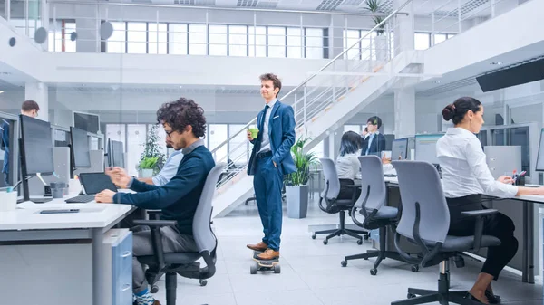 Jonge Happy Handsome Manager draagt een pak en stropdas rijdt een Longboard. Hij houdt een kop koffie vast. Diverse en gemotiveerde zakenmensen werken aan computers in de moderne open kantooromgeving. — Stockfoto