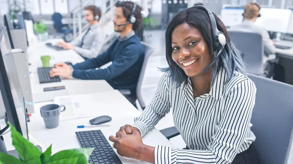 Linda jovem operadora de atendimento ao cliente sorrindo para um retrato em um call center moderno ocupado com equipe multicultural diversificada de especialistas em escritório que usam fones de ouvido e atendem chamadas. — Fotografia de Stock