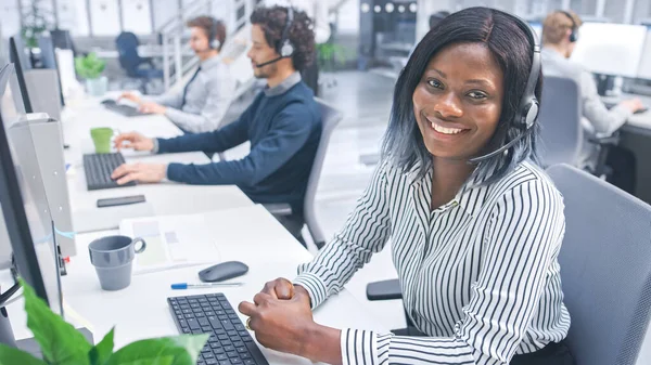 Linda jovem operadora de atendimento ao cliente sorrindo para um retrato em um call center moderno ocupado com equipe multicultural diversificada de especialistas em escritório que usam fones de ouvido e atendem chamadas. — Fotografia de Stock