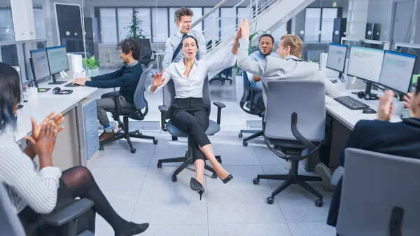 Funcionário alegre empurra sua bela liga feminina em uma cadeira entre linhas de mesas e dar um alto cinco. Diversas pessoas de negócios que trabalham em computadores desktop no espaço de escritório moderno. — Fotografia de Stock