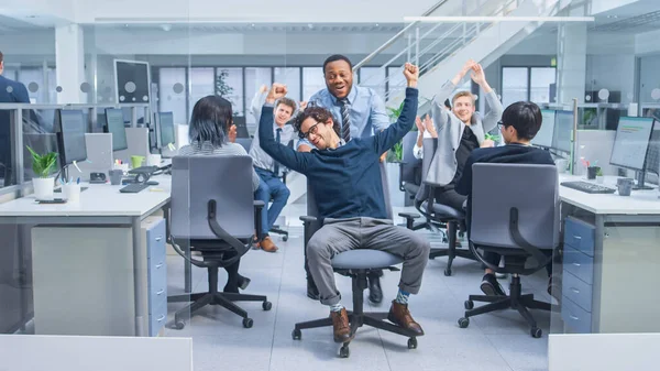 Cheerful Employee mendorong Handsome Colleague-nya pada Kursi Antara Baris-Baris Meja dengan Orang-orang Bisnis Diverse yang bekerja pada Komputer Desktop di Ruang Kantor Modern. — Stok Foto