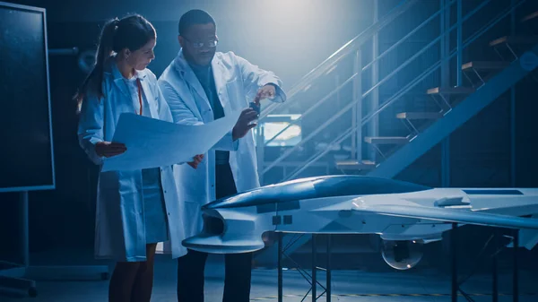 Two Aerospace Engineers Work On Unmanned Aerial Vehicle Drone Prototype. Aviation Scientists in White Coats Holding Blueprints. Laboratory with Commercial Aerial Surveillance Aircraft