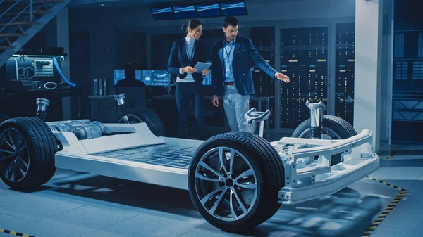 여성 수석 엔지니어는 태블릿 컴퓨터를 사용하며 , 《 중요 한 투자에 대한 자동 전기 자동차 개념 》 (Autonomous Electric Car Concept to Important Investor) 을 소개 한다. 자동차 차대에는 바퀴, 엔진 및 배터리가 있다 — 스톡 사진