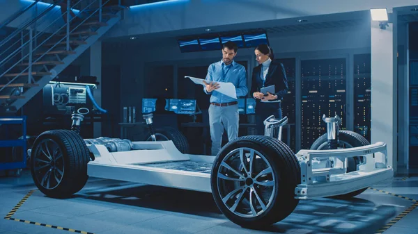 Auto Industry Design Facility: Férfi vezető mérnök mutatja autó tervrajzok Női szoftvertervező és integrációs mérnök. Elektromos jármű platform alváz koncepció kerekek, motor és akkumulátor — Stock Fotó