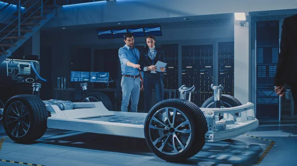 自動車産業デザイン施設:男性チーフエンジニアは女性の車デザイナーに車のプロトタイプを表示します。ホイール、エンジン、バッテリーを搭載した電動車両プラットフォームシャーシのコンセプト. — ストック写真
