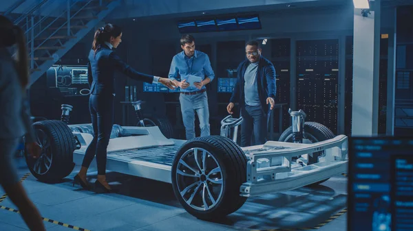 La directora de proyecto habla sobre el prototipo de coche eléctrico con su equipo de ingenieros, mostrando chasis, explicando la sostenibilidad y la eficiencia ecológica del motor — Foto de Stock