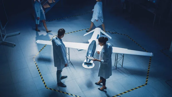 Two Aerospace Engineers Work On Unmanned Aerial Vehicle Drone Prototype. Aviation Scientists in White Coats Talking, Using Tablet Computer. Industrial Laboratory with Surveillance or Military Aircraft