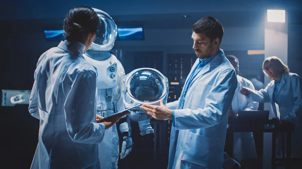 Διαφορετική ομάδα αεροδιαστημικών επιστημόνων και μηχανικών που φορούν λευκά παλτά έχουν συζήτηση, χρησιμοποιούν υπολογιστές, κατασκευάζουν Astronaut κράνος για νέα διαστημική στολή προσαρμοσμένη για Galaxy Exploration και Travel. — Φωτογραφία Αρχείου