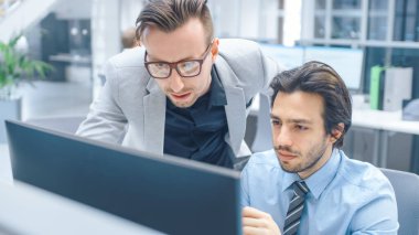 Parlak Modern Ofis: Yakışıklı Ofis İşçisi Masaüstü Bilgisayarında Oturup Çalışıyor, Proje Yöneticisi ile Sorun Çözümünü Tartışıyor. İş Adamları Planlama ve Problem Çözme Ekibi
