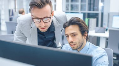 Parlak Modern Ofis: Yakışıklı Ofis İşçisi Masaüstü Bilgisayarında Oturup Çalışıyor, Proje Yöneticisi ile Sorun Çözümünü Tartışıyor. İş Adamları Planlama ve Problem Çözme Ekibi