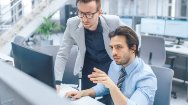 Parlak Modern Ofis: Yakışıklı Ofis İşçisi Masaüstü Bilgisayarında Oturup Çalışıyor, Proje Yöneticisi ile Sorun Çözümünü Tartışıyor. İş Adamları Planlama ve Problem Çözme Ekibi