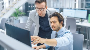 Parlak Modern Ofis: Yakışıklı Ofis İşçisi Masaüstü Bilgisayarında Oturup Çalışıyor, Proje Yöneticisi ile Sorun Çözümünü Tartışıyor. İş Adamları Planlama ve Problem Çözme Ekibi