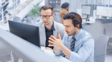 Parlak Modern Ofis: Yakışıklı Ofis İşçisi Masaüstü Bilgisayarında Oturup Çalışıyor, Erkek Proje Yöneticisi ile Sorun Çözümünü Tartışıyor. Çeşitli İş Adamları Ekibi Sorunları Çözüyor